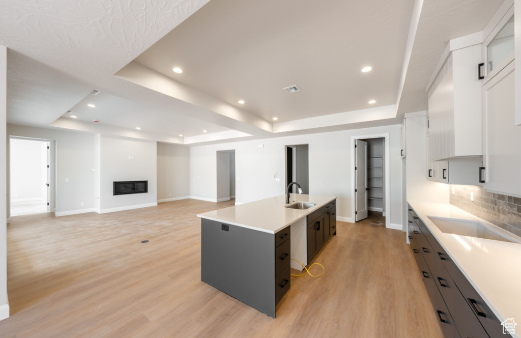 Kitchen with a tray ceiling, a kitchen island with sink, light countertops, recessed lighting, and a glass covered fireplace