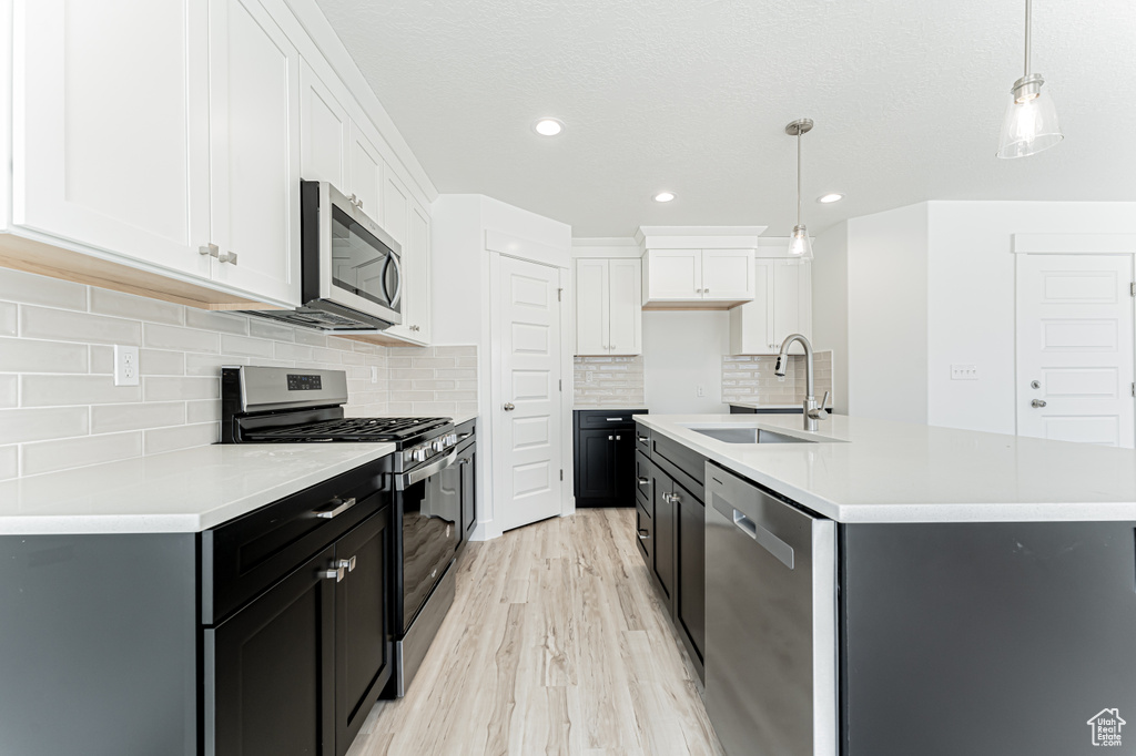Kitchen featuring appliances with stainless steel finishes, white cabinetry, light wood-style floors, pendant lighting, and an island with sink