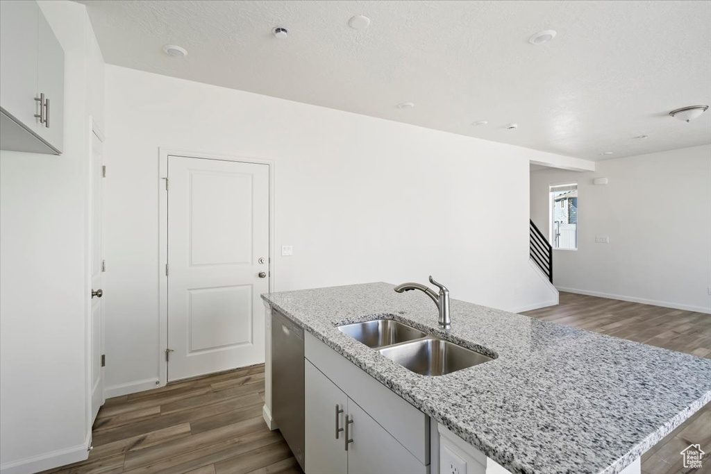 Kitchen featuring light stone counters, wood finished floors, a sink, stainless steel dishwasher, and an island with sink