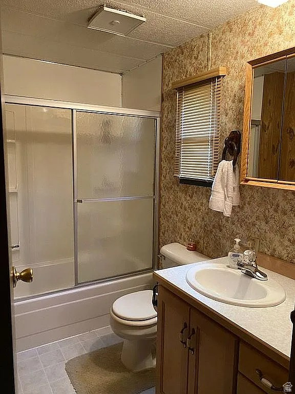 Full bath with wallpapered walls, bath / shower combo with glass door, toilet, tile patterned flooring, and vanity
