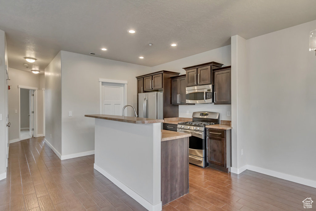 Kitchen featuring dark brown cabinetry, recessed lighting, stainless steel appliances, wood tiled floor, and an island with sink