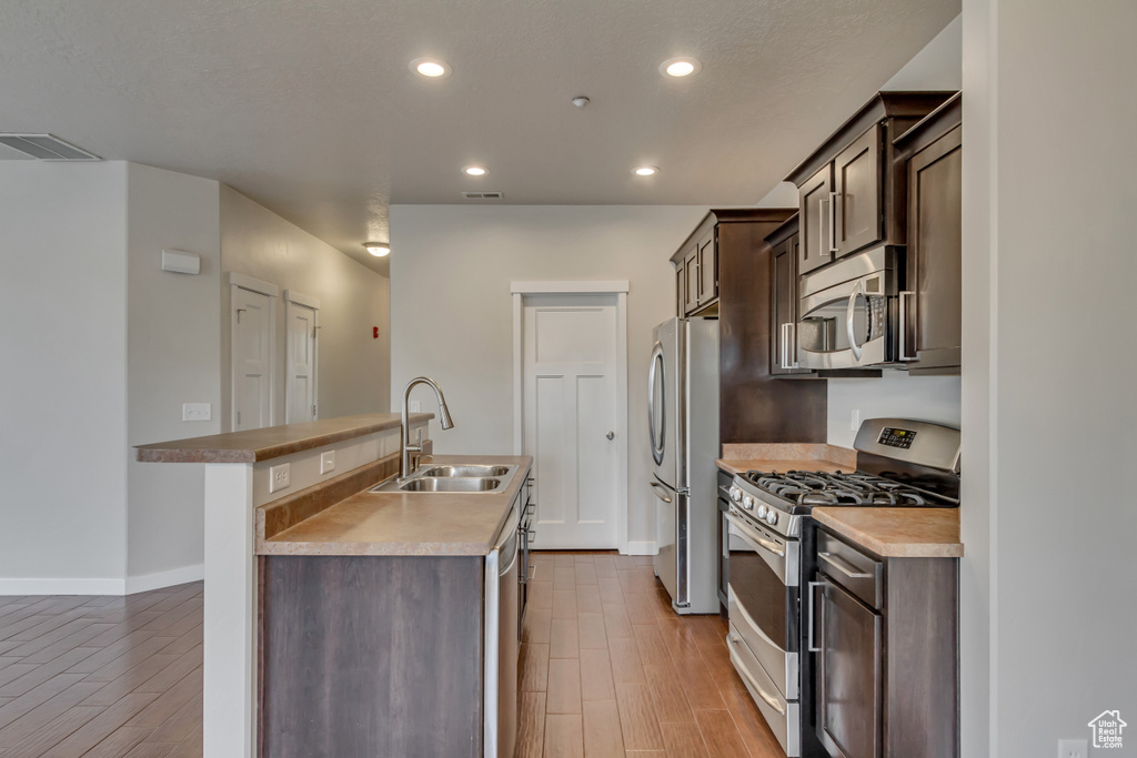 Kitchen featuring a center island with sink, stainless steel appliances, dark brown cabinetry, a sink, and wood finished floors