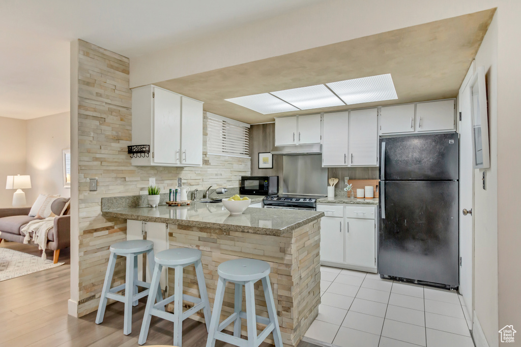 Kitchen with light tile patterned floors, a peninsula, under cabinet range hood, black appliances, and white cabinetry