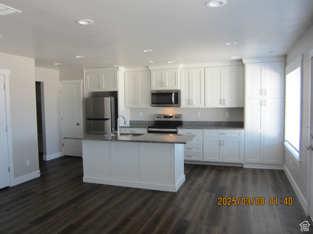 Kitchen featuring dark wood-style floors, appliances with stainless steel finishes, white cabinets, a sink, and an island with sink