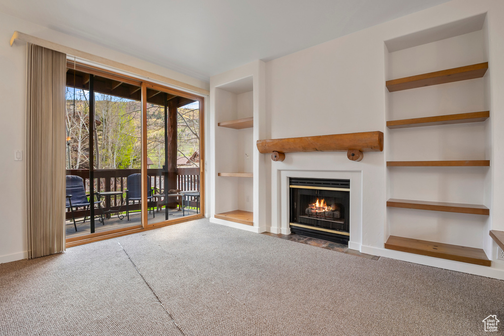 Unfurnished living room featuring carpet floors, built in shelves, a fireplace with flush hearth, and baseboards