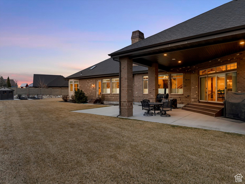 Back of property at dusk with a lawn, entry steps, a patio, brick siding, and a chimney