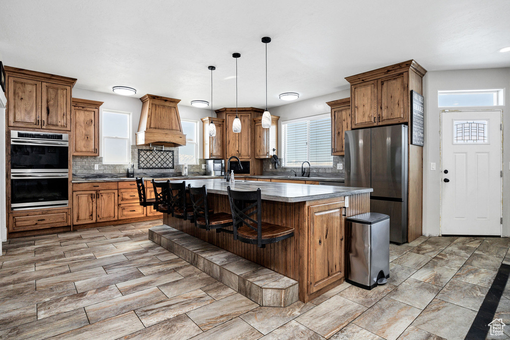 Kitchen with premium range hood, a kitchen bar, a kitchen island with sink, stainless steel appliances, and brown cabinetry