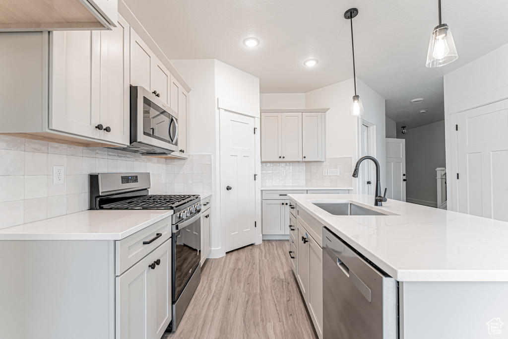 Kitchen featuring appliances with stainless steel finishes, backsplash, a center island with sink, light wood finished floors, and decorative light fixtures
