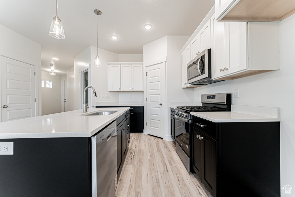 Kitchen featuring stainless steel appliances, dark cabinets, a center island with sink, light wood-type flooring, and light countertops