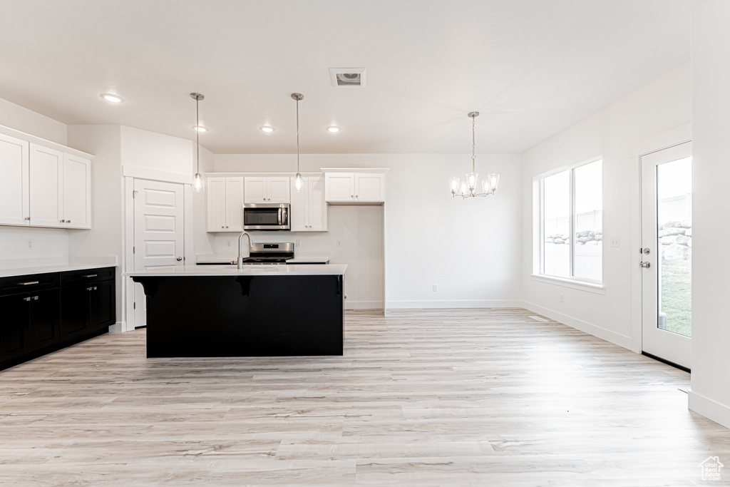Kitchen featuring appliances with stainless steel finishes, light wood-style floors, a kitchen island with sink, white cabinets, and decorative light fixtures