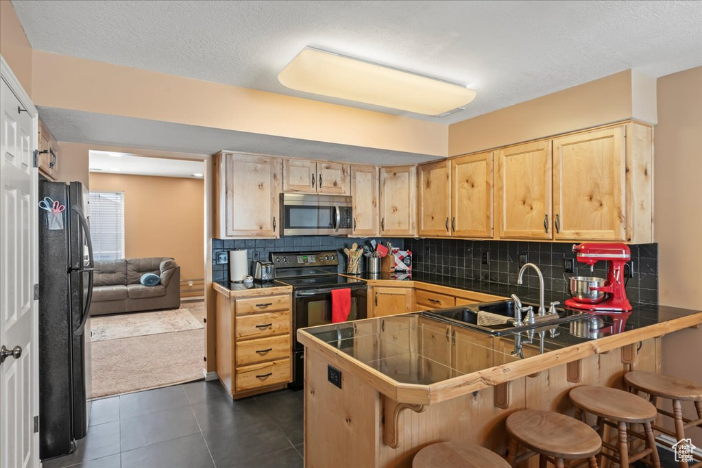 Kitchen with a breakfast bar area, a peninsula, light brown cabinetry, backsplash, and black appliances