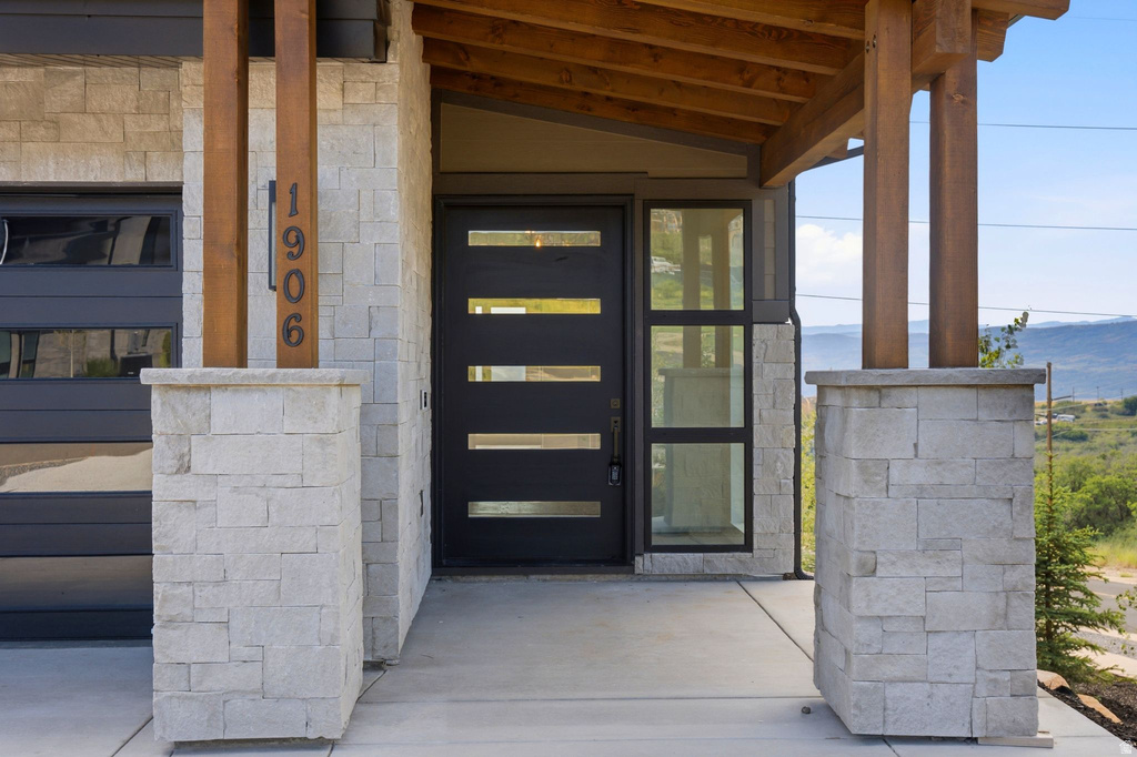 Contemporary entry featuring a dark-finish door with horizontal glass panels, stone veneer siding, wood beams, and an exposed aggregate concrete walkway