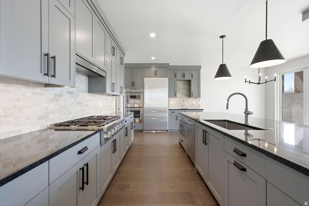 Gourmet kitchen featuring extensive light gray cabinetry, dark countertops, a large center island with an undermount sink, stainless steel appliances, and wood-finish flooring