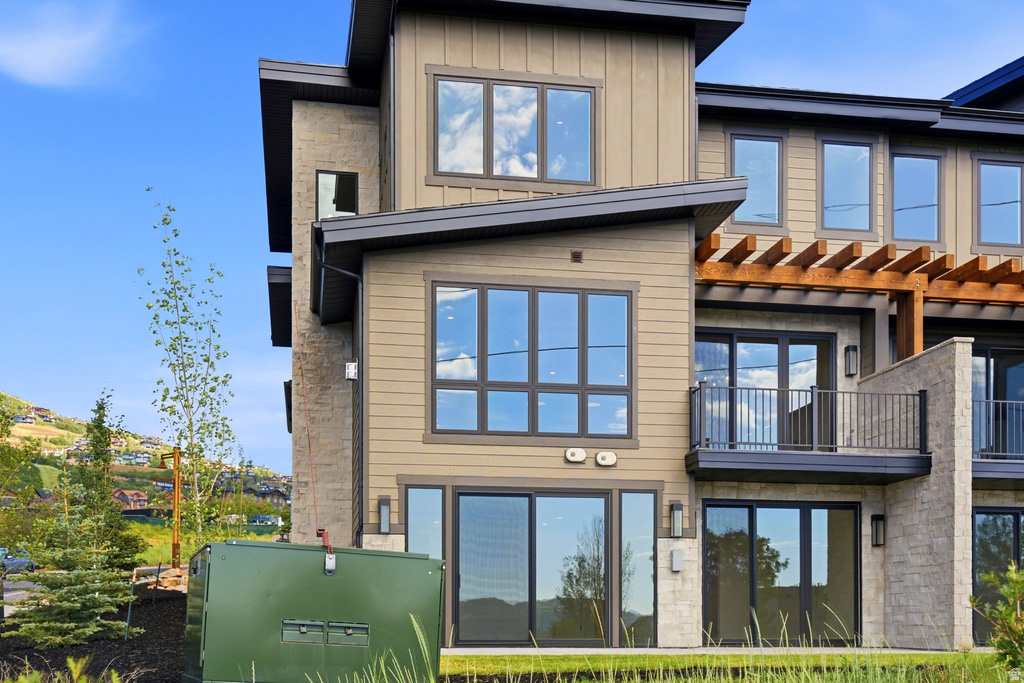 Modern multi-level exterior featuring tan vertical siding, stone accents, multiple large windows, and a private balcony with metal railings