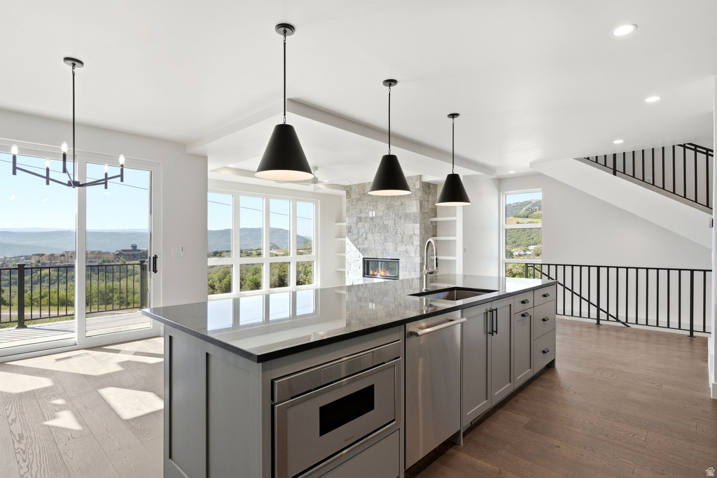 Open-concept living space featuring an expansive kitchen island with a dark countertop and integrated stainless steel appliances, wood-finish flooring, a contemporary fireplace with a stone surround, and large windows framing the outdoor scenery