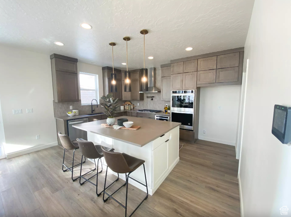 Kitchen with wall chimney range hood, decorative backsplash, light wood-style floors, stainless steel appliances, and a sink