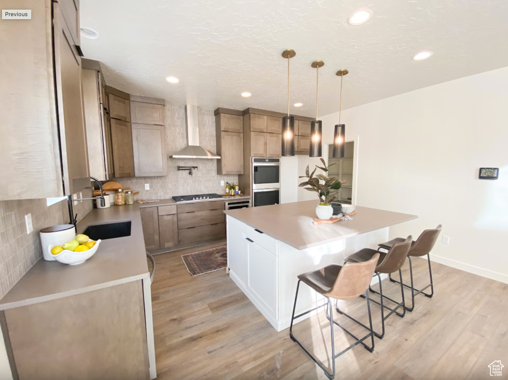 Kitchen featuring tasteful backsplash, stainless steel appliances, light wood-style floors, a breakfast bar area, and wall chimney range hood