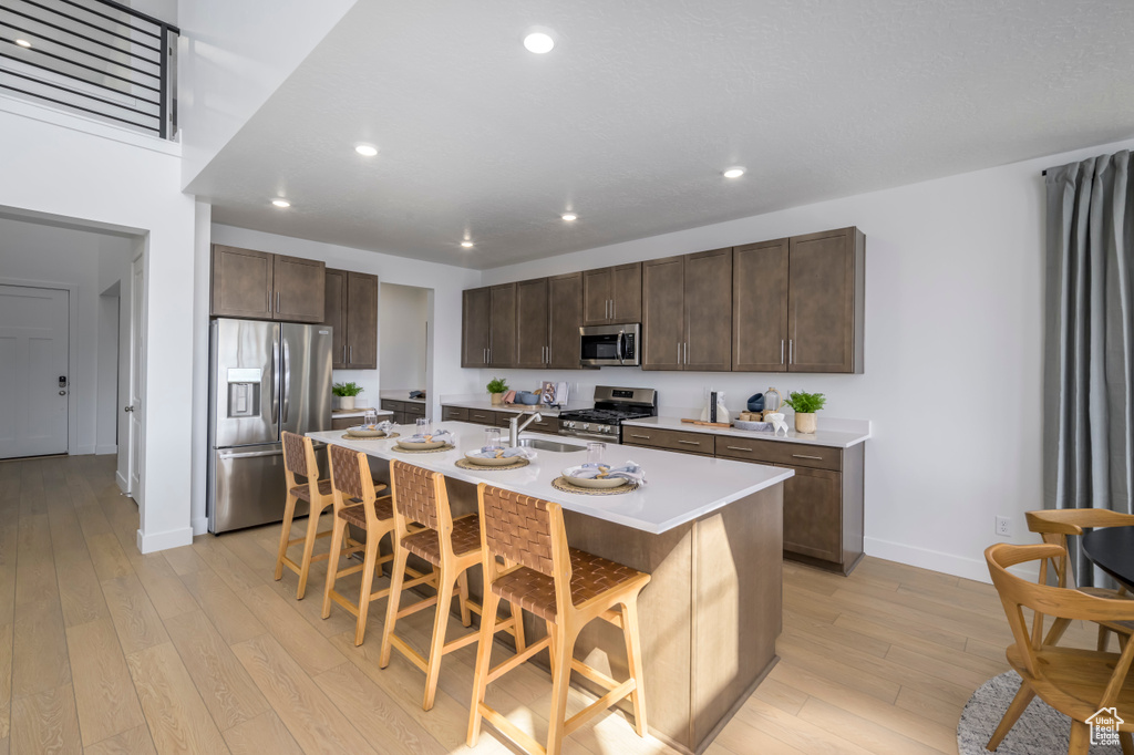 Kitchen featuring light wood-type flooring, an island with sink, stainless steel appliances, a breakfast bar area, and dark brown cabinets
