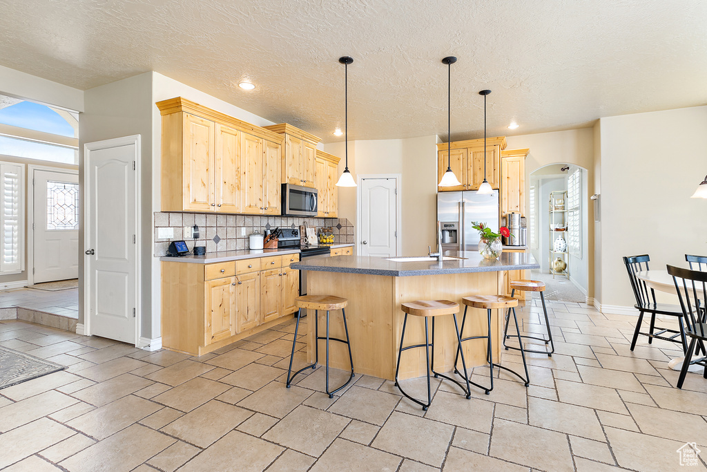 Kitchen featuring light brown cabinets, arched walkways, stainless steel appliances, decorative backsplash, and a kitchen bar