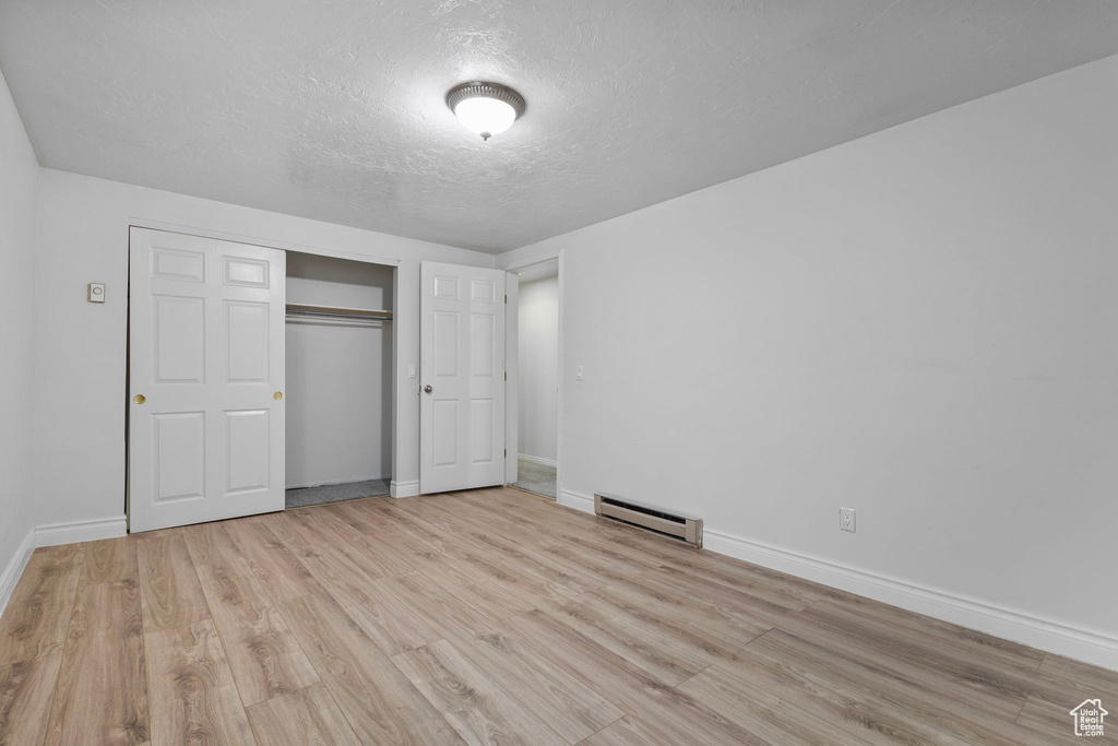 Unfurnished bedroom with light wood-style flooring, a textured ceiling, a closet, a baseboard radiator, and baseboards