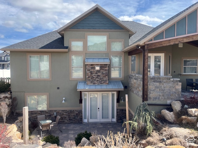 Back of property with a shingled roof, stucco siding, french doors, stone siding, and a patio