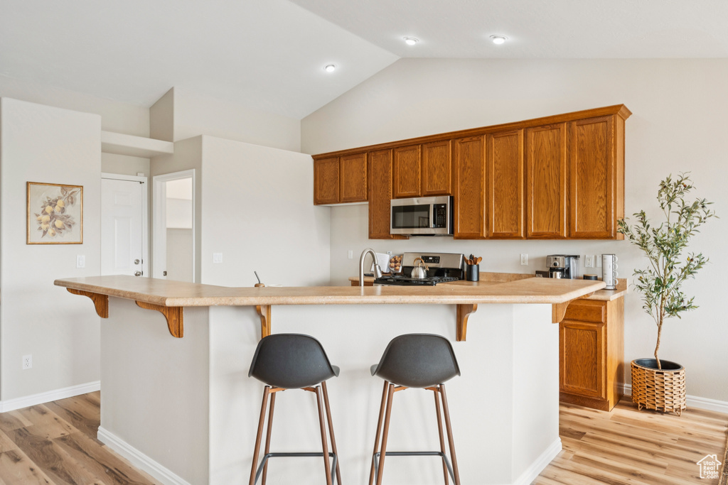 Kitchen with stainless steel appliances, brown cabinetry, a kitchen breakfast bar, light wood finished floors, and vaulted ceiling
