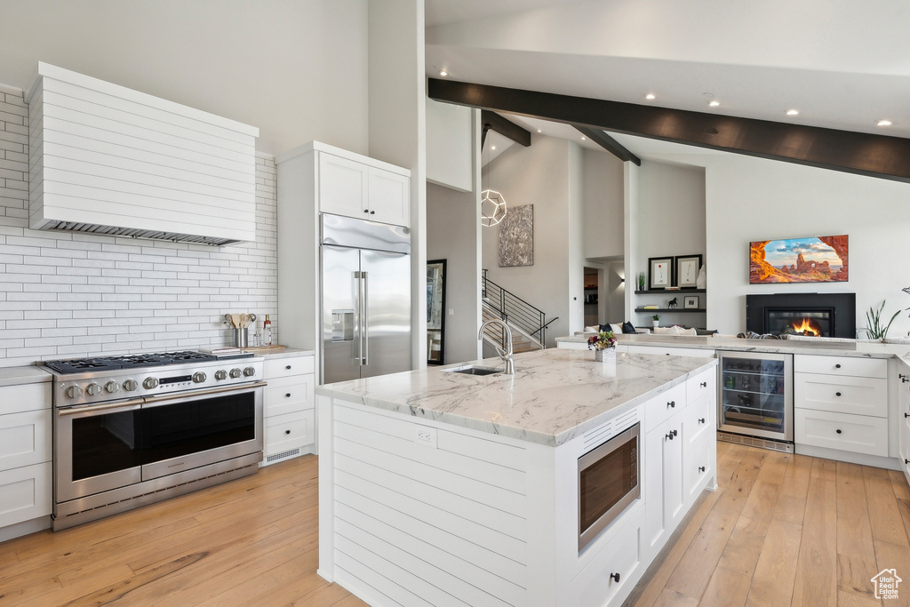 Kitchen featuring a center island with sink, beam ceiling, wine cooler, built in appliances, and custom range hood