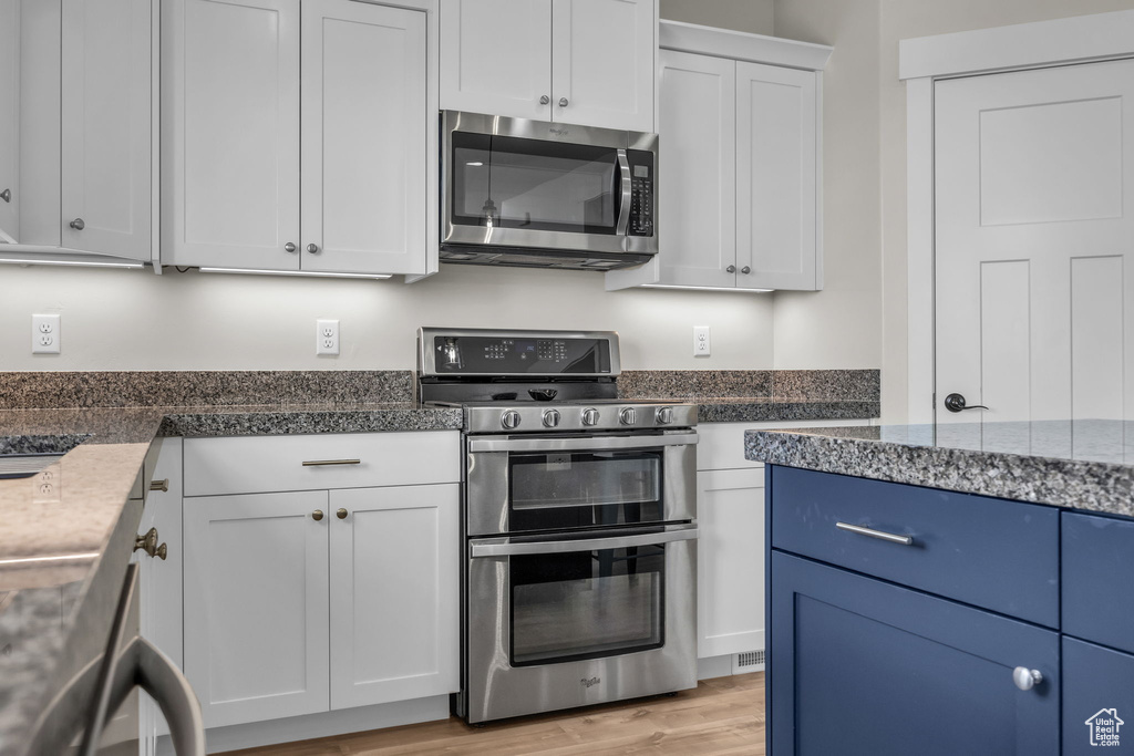 Kitchen with dark stone countertops, light wood finished floors, blue cabinetry, white cabinets, and appliances with stainless steel finishes