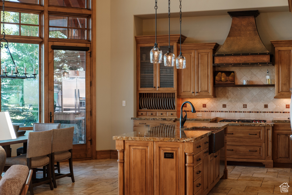 Kitchen featuring custom range hood, stone tile flooring, light stone counters, stainless steel gas stovetop, and decorative backsplash