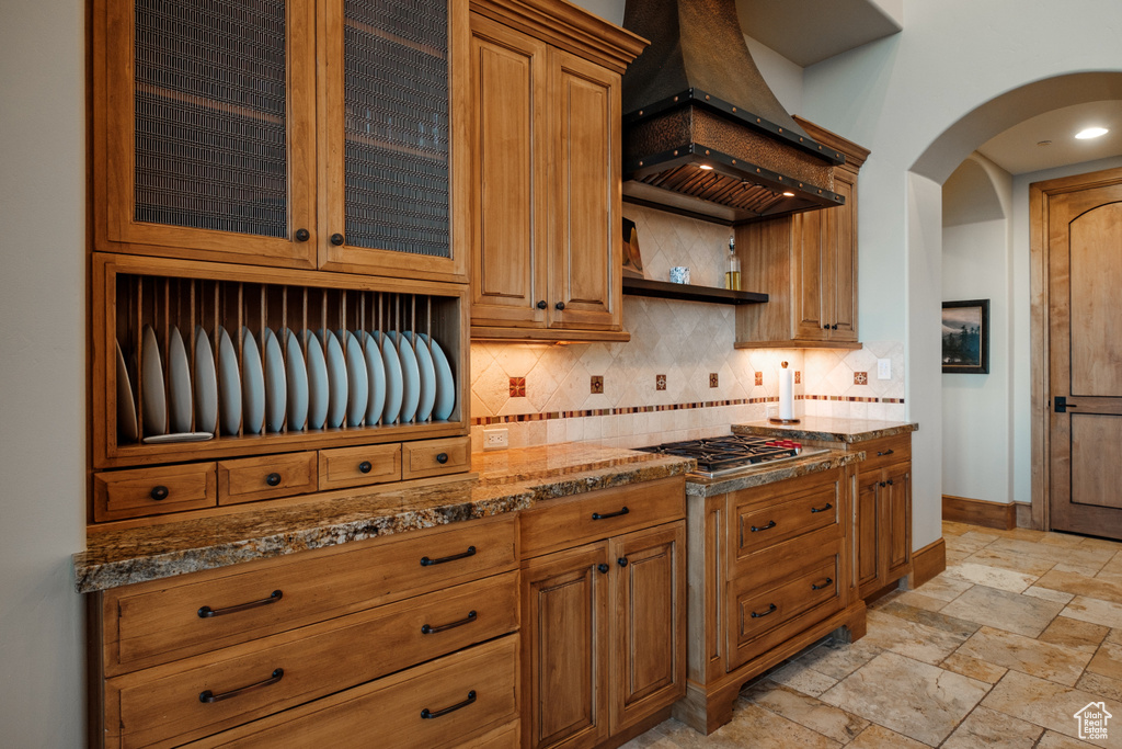 Kitchen with stainless steel gas cooktop, custom range hood, backsplash, arched walkways, and dark stone countertops