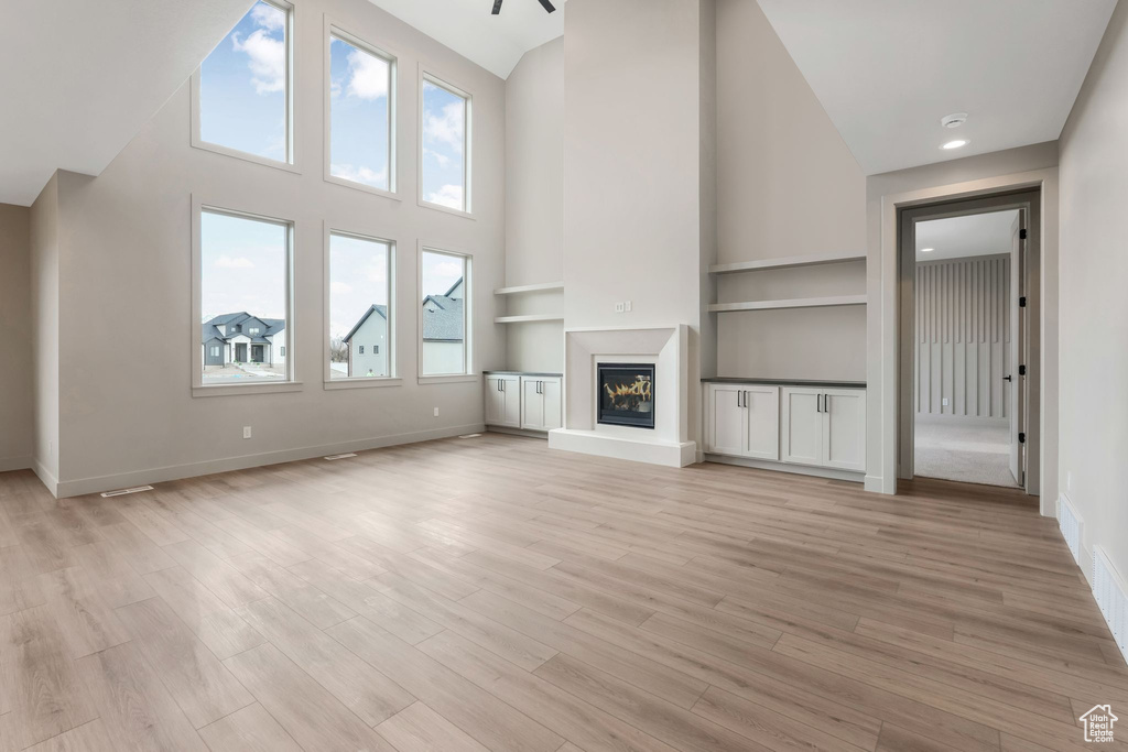 Unfurnished living room featuring light wood-type flooring, built in shelves, a glass covered fireplace, baseboards, and a towering ceiling