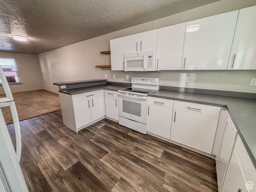 Kitchen with open shelves, dark countertops, dark wood-style floors, white appliances, and a peninsula