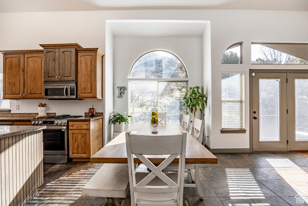 Kitchen featuring stainless steel appliances, dark stone counters, stone tile floors, brown cabinets, and baseboards