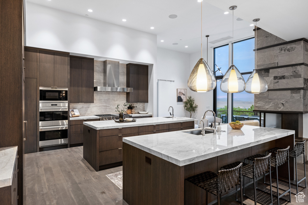 Kitchen featuring a sink, a spacious island, modern cabinets, stainless steel appliances, and wall chimney range hood