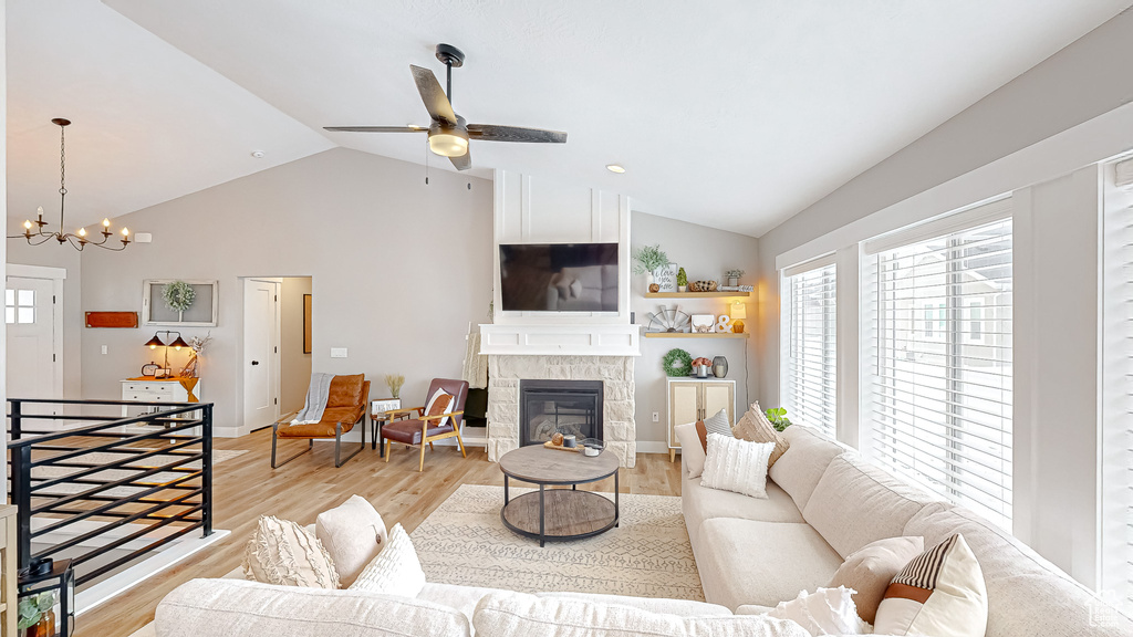 Living room with lofted ceiling, light wood-style flooring, ceiling fan with notable chandelier, a stone fireplace, and baseboards