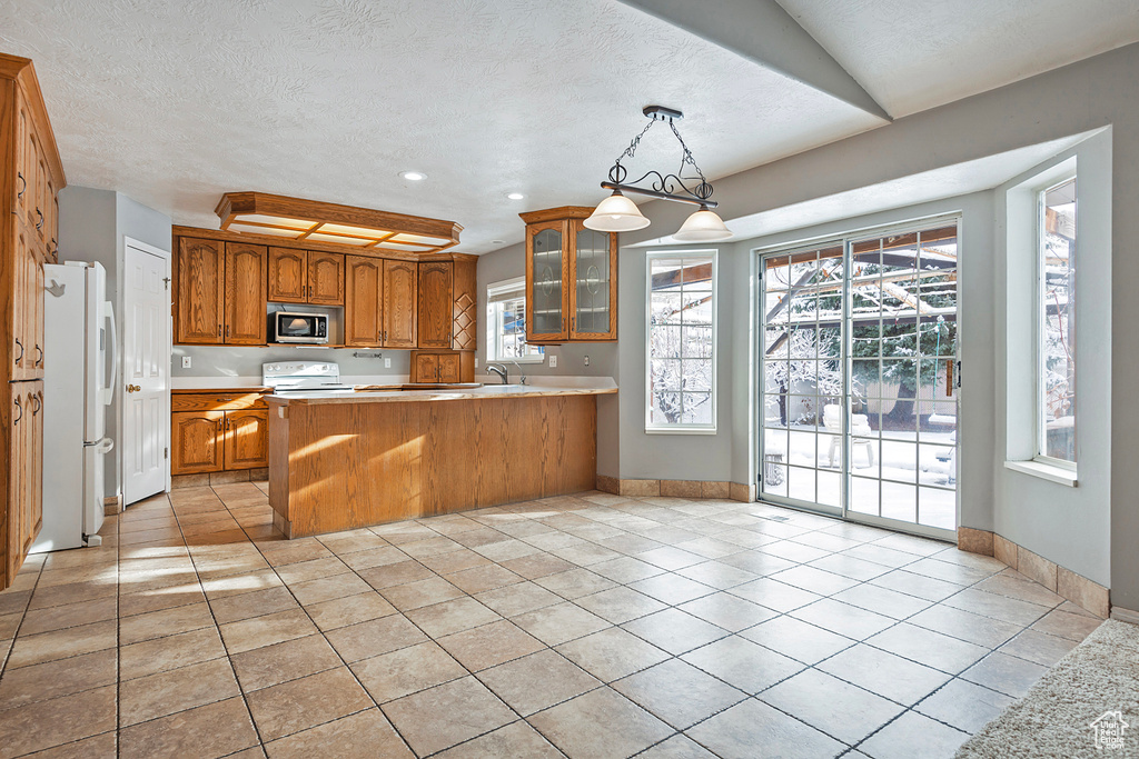 Kitchen with white appliances, a peninsula, brown cabinetry, light countertops, and glass insert cabinets