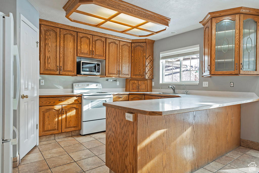 Kitchen featuring brown cabinets, a sink, white appliances, a peninsula, and glass insert cabinets