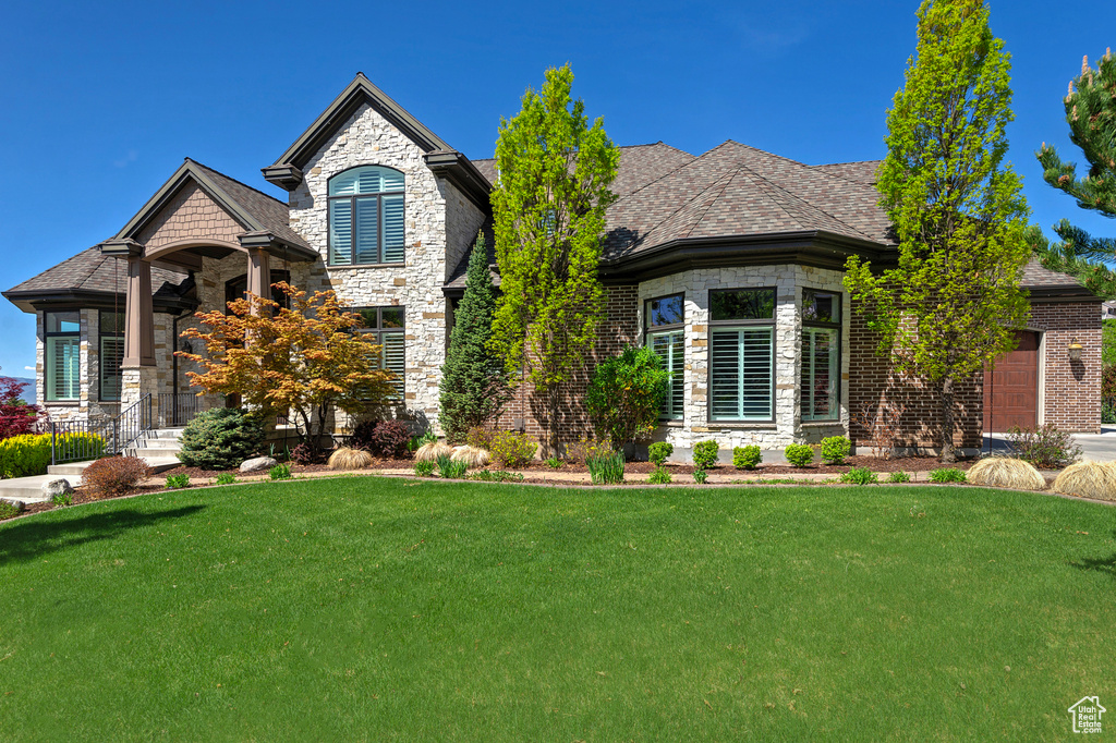 French country inspired facade with a garage, stone siding, a front yard, and a shingled roof