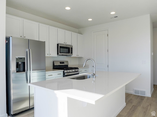 Kitchen featuring stainless steel appliances, a sink, white cabinets, an island with sink, and light wood-type flooring