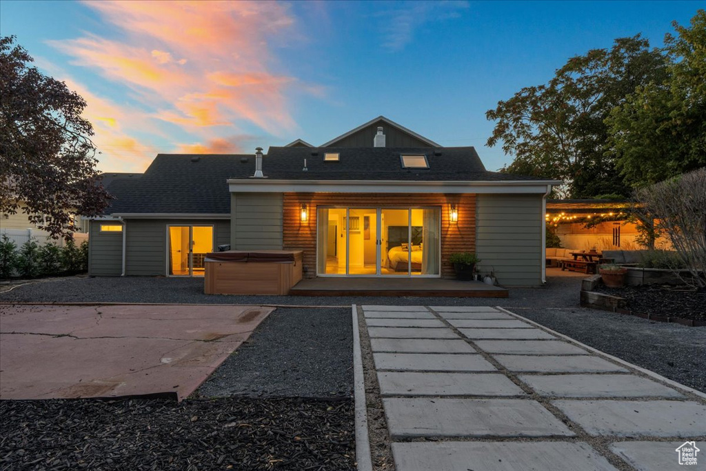 Back of house at dusk featuring a hot tub, a patio, and a shingled roof