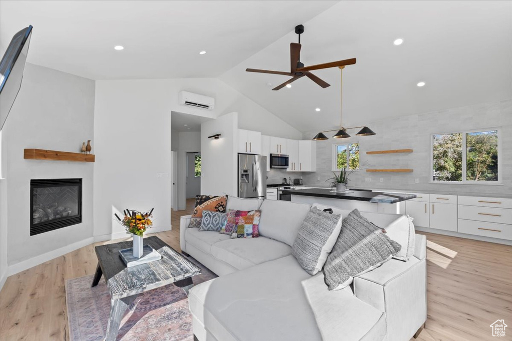 Living room with high vaulted ceiling, a wall mounted AC, ceiling fan, a glass covered fireplace, and light wood-type flooring