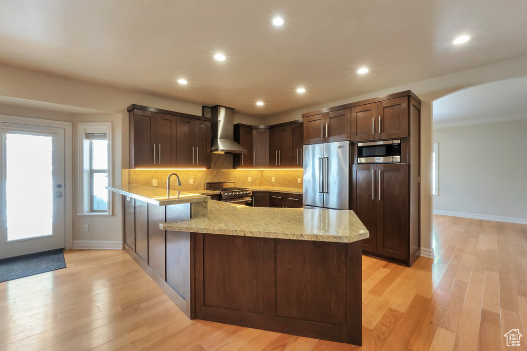 Kitchen with high end appliances, wall chimney range hood, a peninsula, dark brown cabinetry, and baseboards