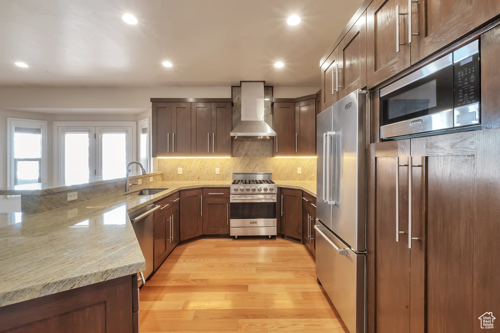 Kitchen featuring high end appliances, wall chimney exhaust hood, a sink, light wood-style flooring, and a peninsula