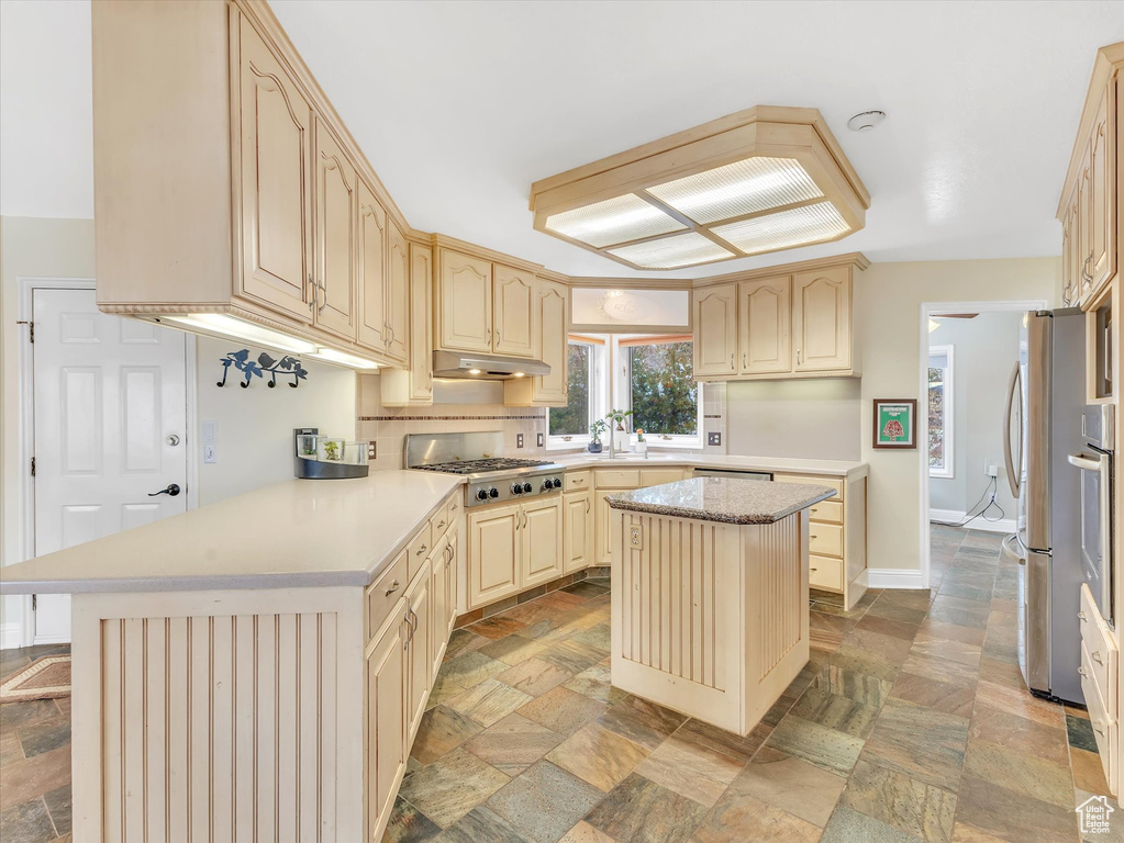 Kitchen with stone tile floors, a peninsula, light countertops, under cabinet range hood, and appliances with stainless steel finishes