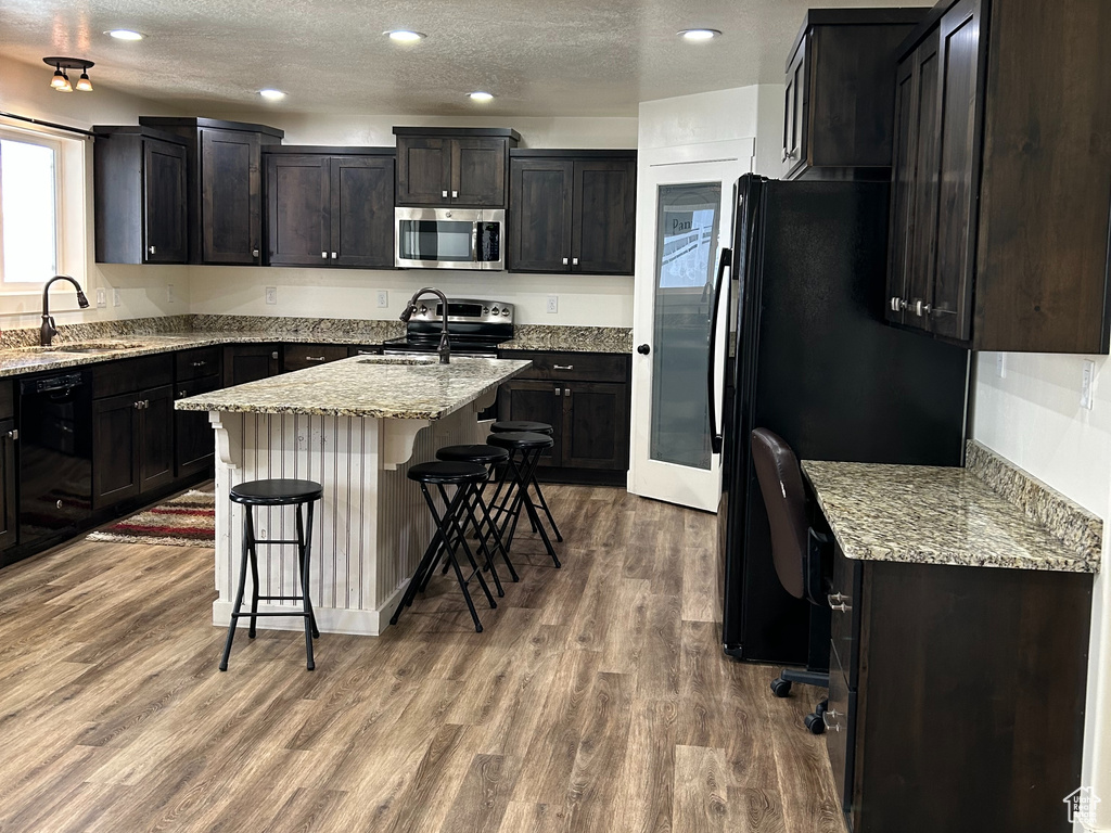 Kitchen with dark wood finished floors, a kitchen bar, light stone counters, black appliances, and a kitchen island with sink