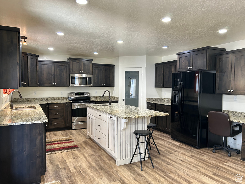 Kitchen with light wood finished floors, a sink, light stone counters, stainless steel appliances, and a kitchen island with sink
