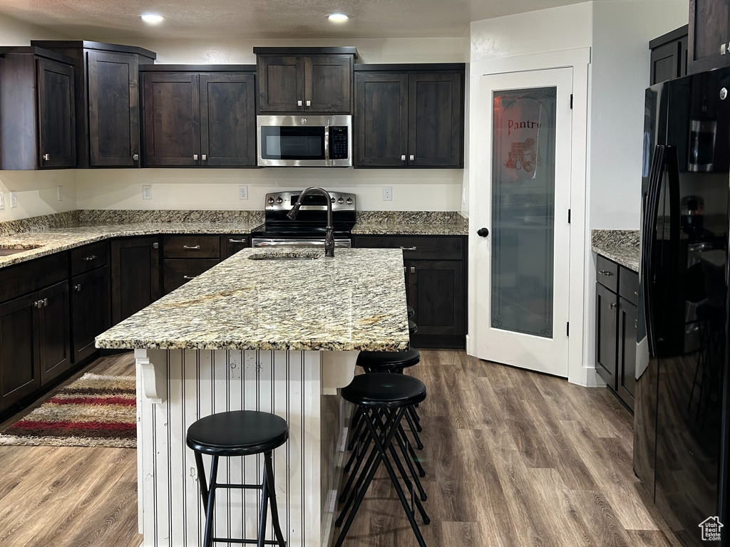 Kitchen with a center island with sink, a breakfast bar area, light stone counters, appliances with stainless steel finishes, and wood finished floors