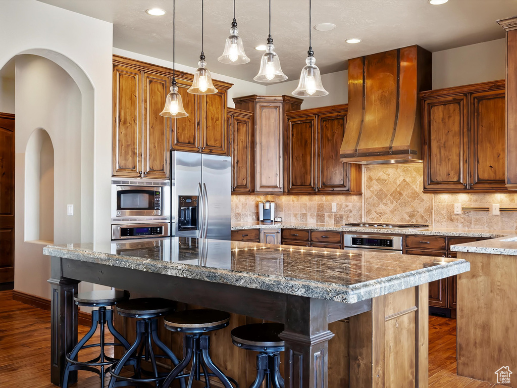 Kitchen with dark wood-type flooring, a breakfast bar area, decorative backsplash, appliances with stainless steel finishes, and custom exhaust hood