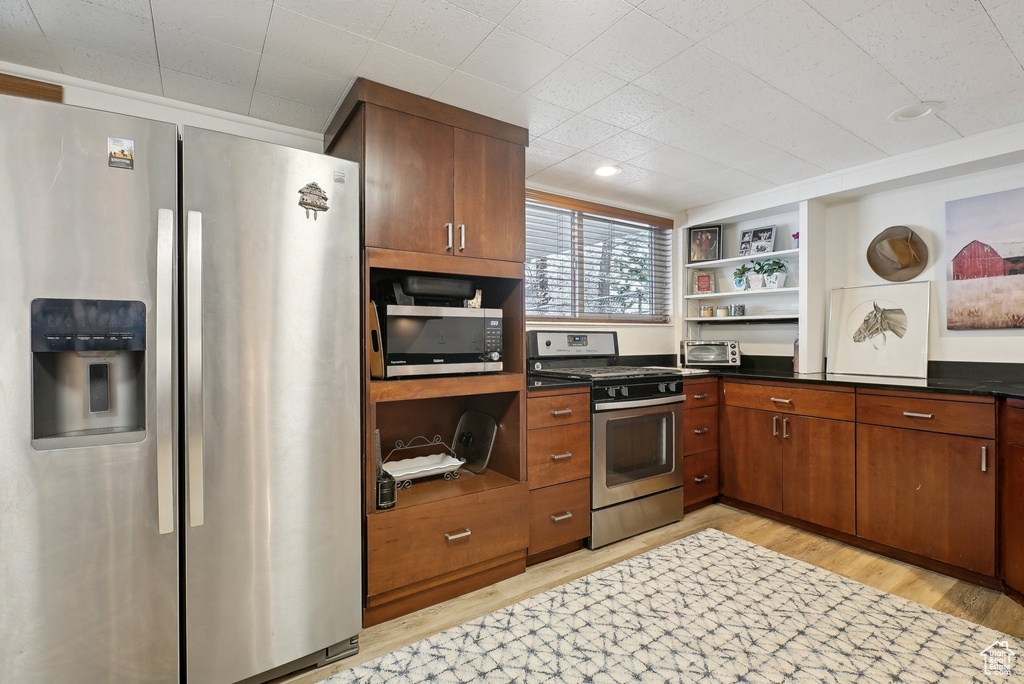 Kitchen with stainless steel appliances, open shelves, light wood-style flooring, dark countertops, and brown cabinetry