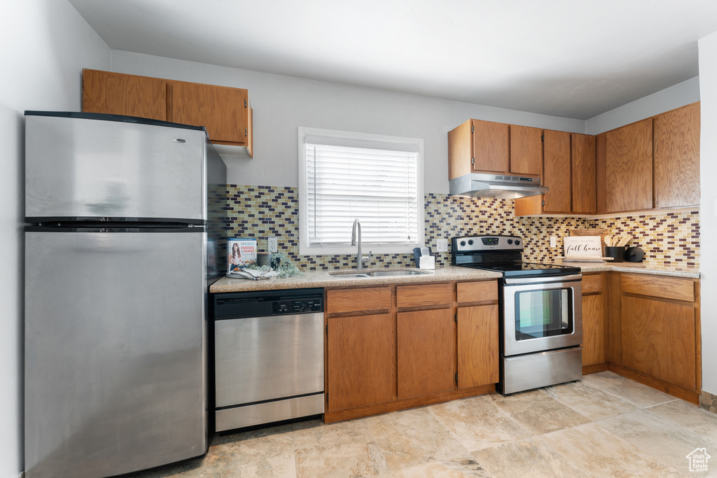 Kitchen featuring tasteful backsplash, under cabinet range hood, brown cabinets, appliances with stainless steel finishes, and a sink
